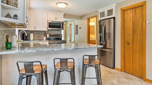 A kitchen with white cabinets, a granite countertop, a stainless steel refrigerator, and two bar stools.