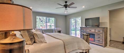 A bedroom with a bed, a ceiling fan, a television, a fireplace, and large windows.