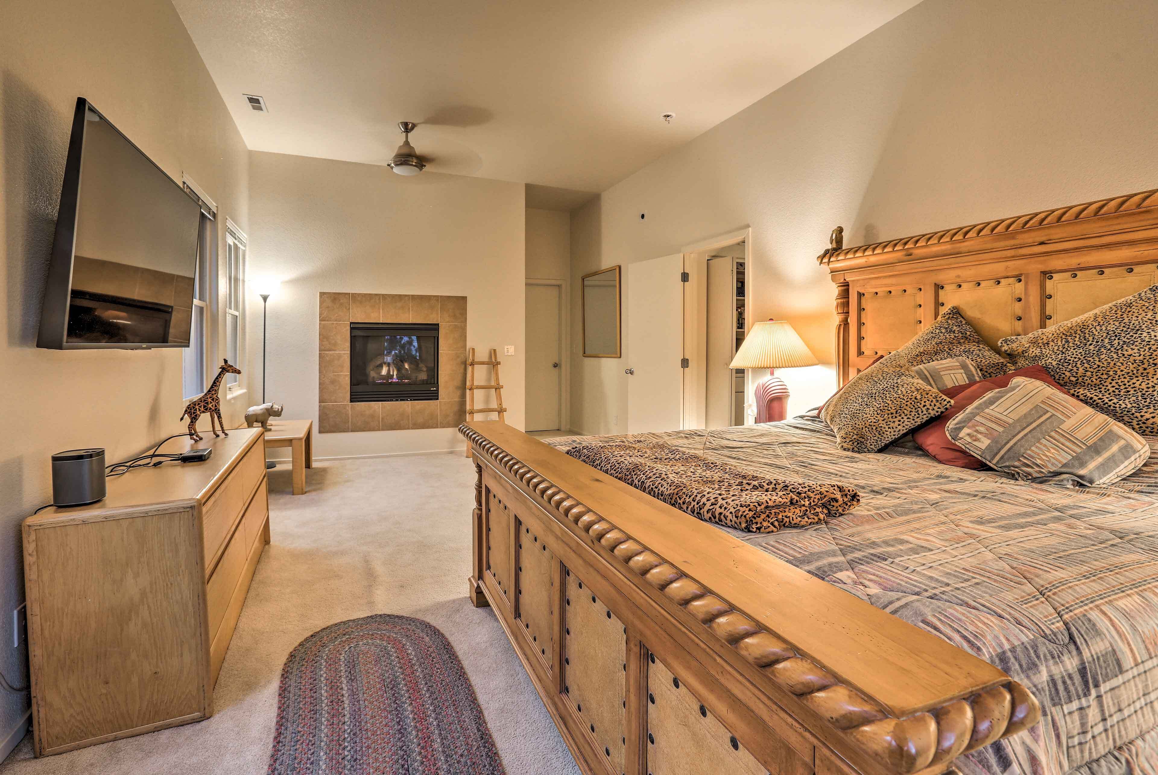 A bedroom with a wooden bed, a television mounted on the wall, a fireplace, and a desk with a lamp.