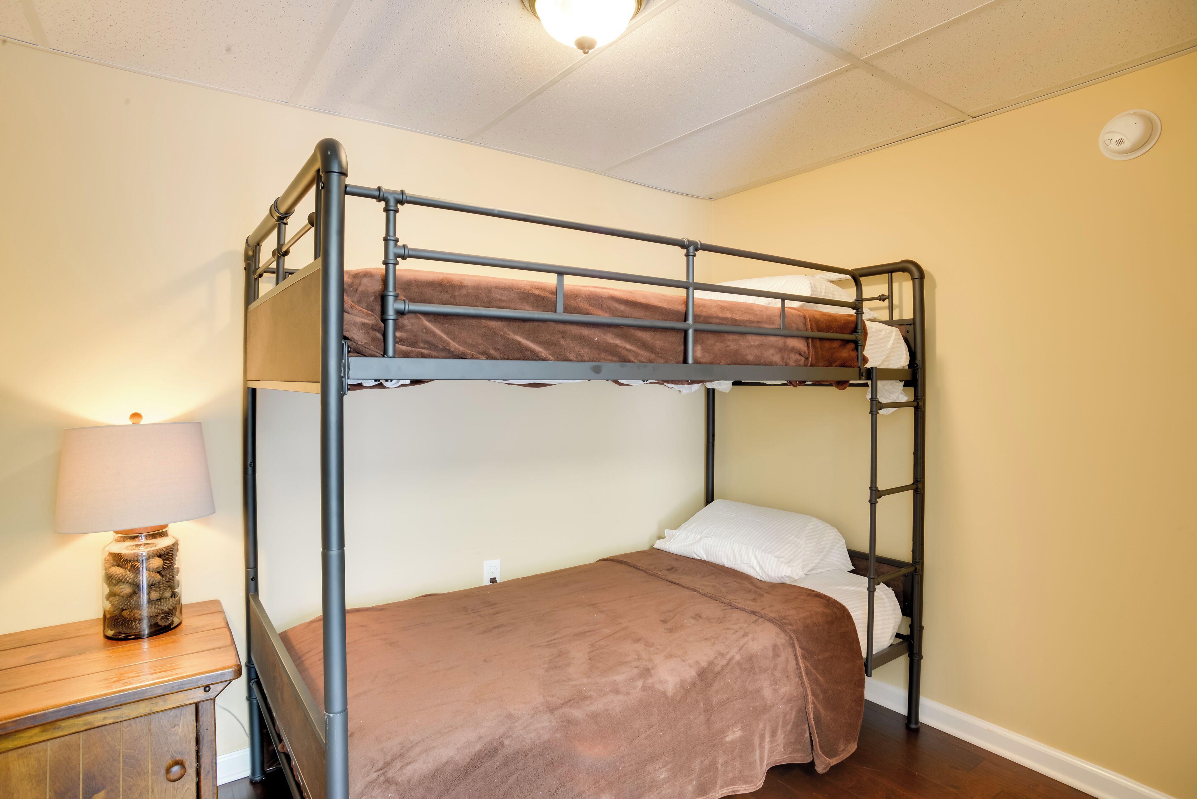 A bunk bed with a brown blanket, a wooden nightstand with a lamp, and a wall socket.