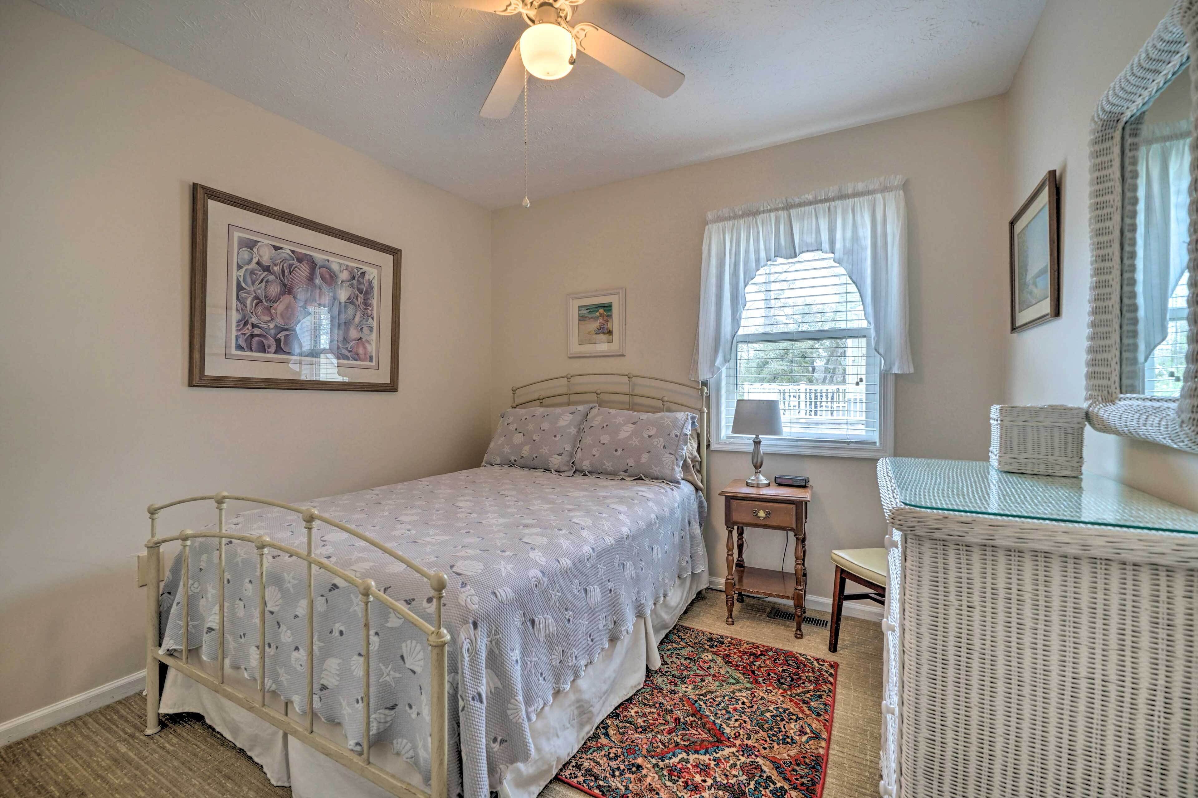 A bedroom with a bed, a nightstand, a wicker side table, a ceiling fan, and a window with curtains.