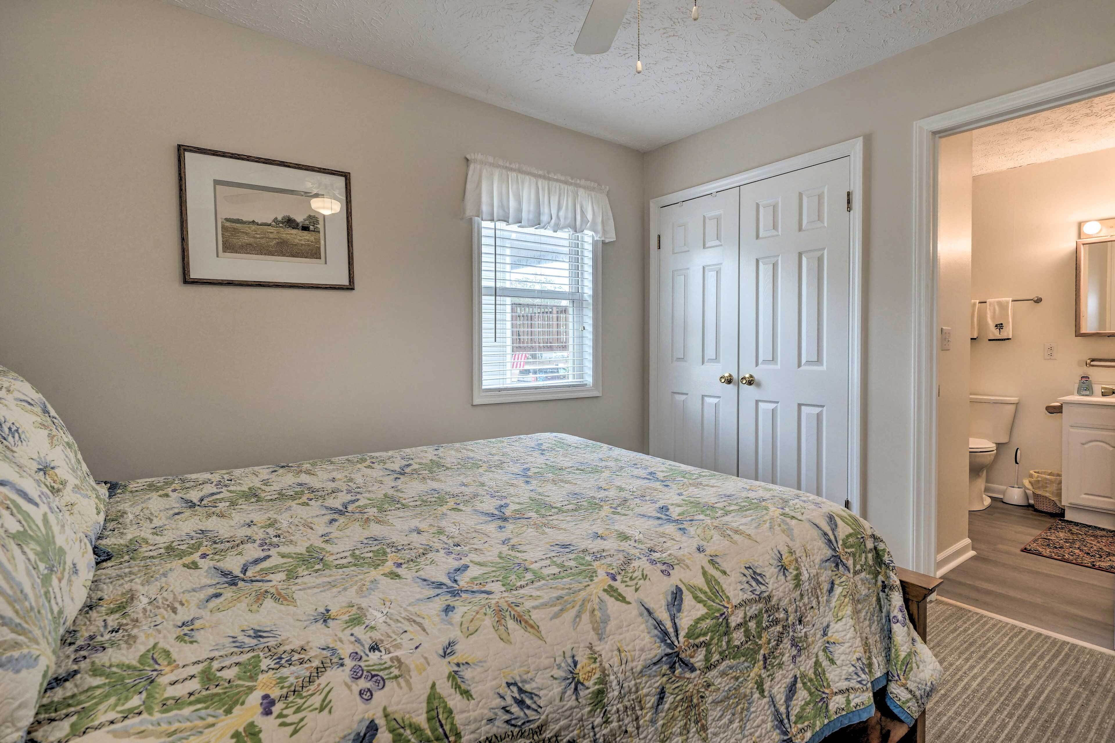 Un dormitorio con cubrecama estampado floral, un cuadro enmarcado en la pared, una ventana con persianas y una puerta que da a un baño.