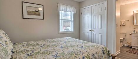 Un dormitorio con cubrecama estampado floral, un cuadro enmarcado en la pared, una ventana con persianas y una puerta que da a un baño.