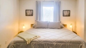 A bedroom with a large bed, two bedside tables, a window with curtains, and two wall-mounted picture frames. - Cozy Bryson City Home w/ Fire Pit & Mtn Views! (Bryson City)
