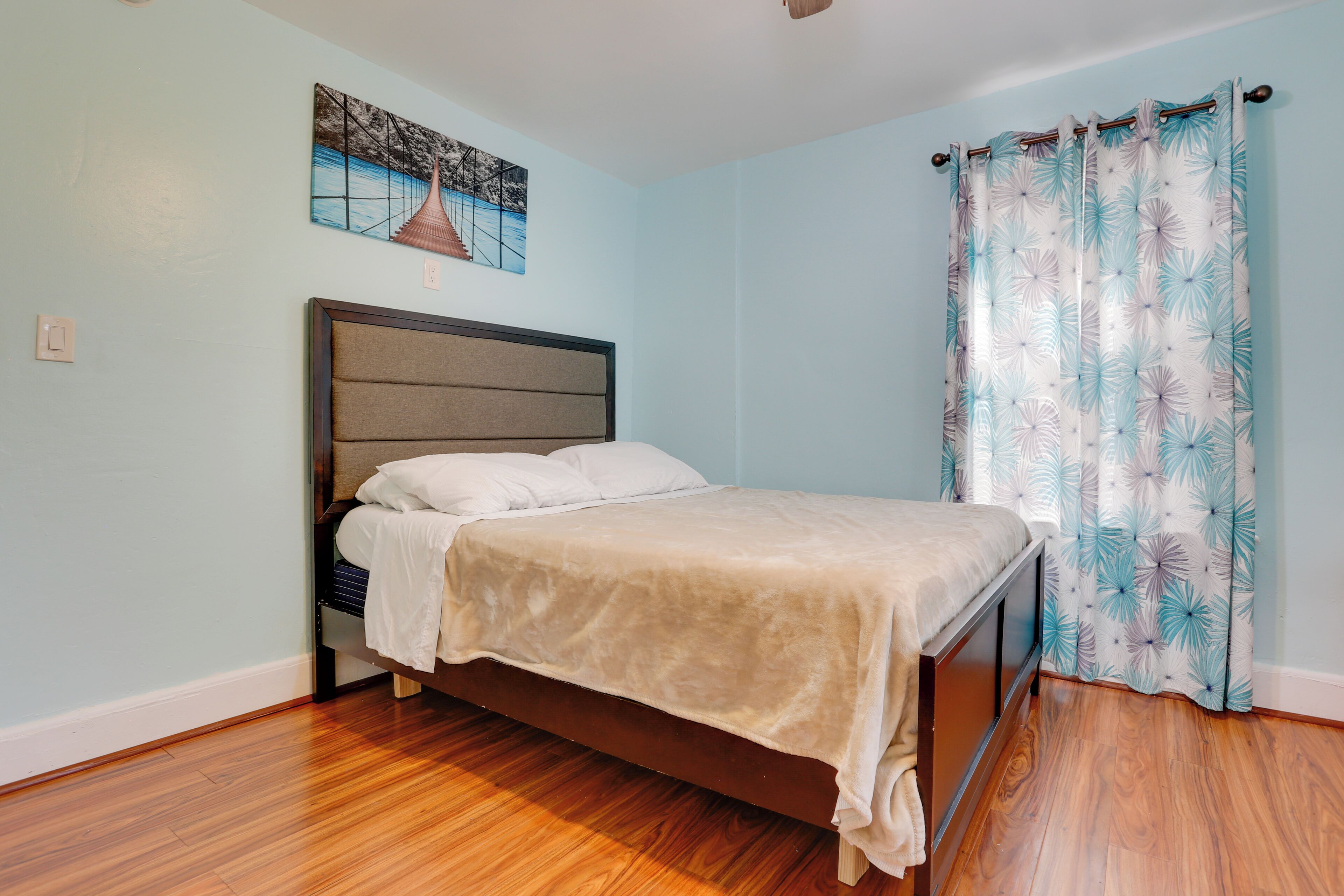 A bedroom with a bed, wooden flooring, a headboard, a painting on the wall, and a curtain with a leaf pattern.