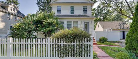 A two-story house with a white picket fence, a front yard with a small garden, and a visible garage door.