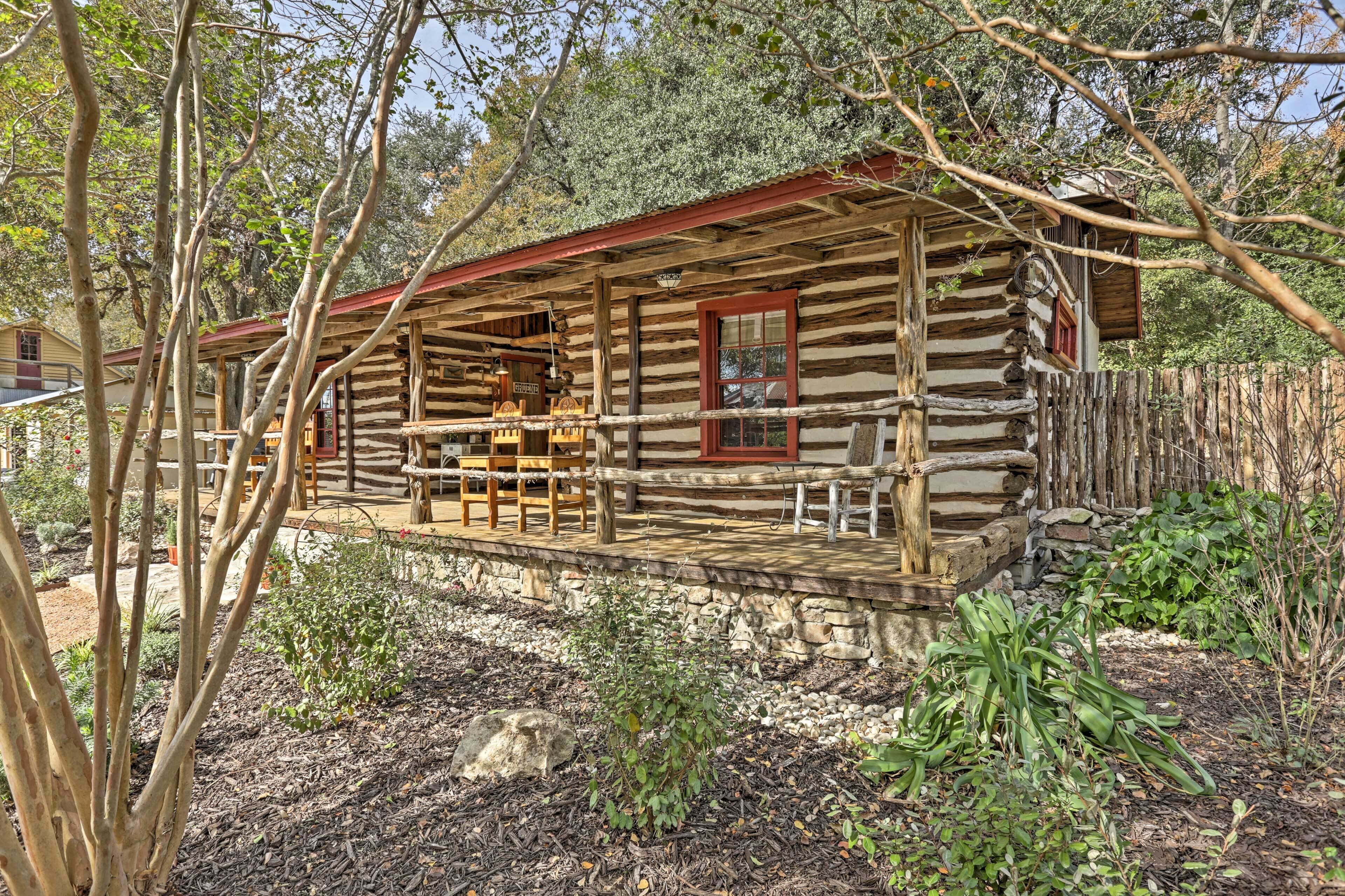 A rustic log cabin with a porch and wooden chairs, surrounded by trees and a stone wall.