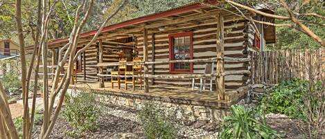 A rustic log cabin with a porch and wooden chairs, surrounded by trees and a stone wall.