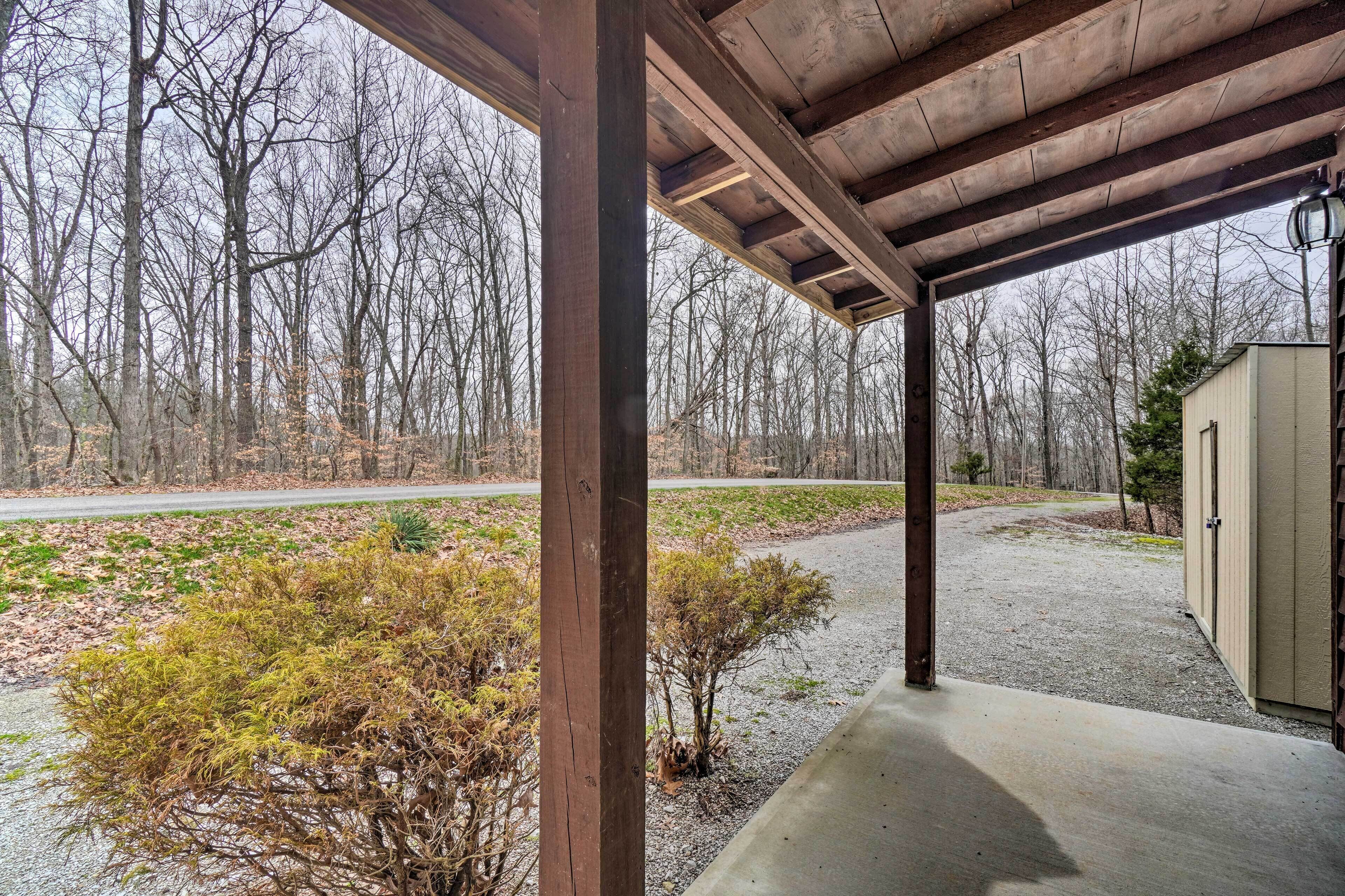 Un espacio exterior techado con suelo de hormigón, vigas de madera y vistas a una zona boscosa.