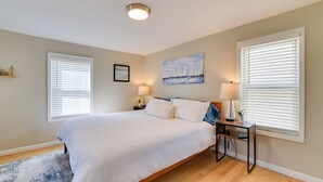 A bedroom with a large bed, two bedside tables, a nightstand, a painting, and a window with blinds.