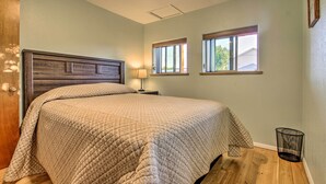 Un dormitorio con cabecera de madera, cama con colcha acolchada, dos ventanas con persianas, una mesita de noche con lámpara y un cesto de basura.