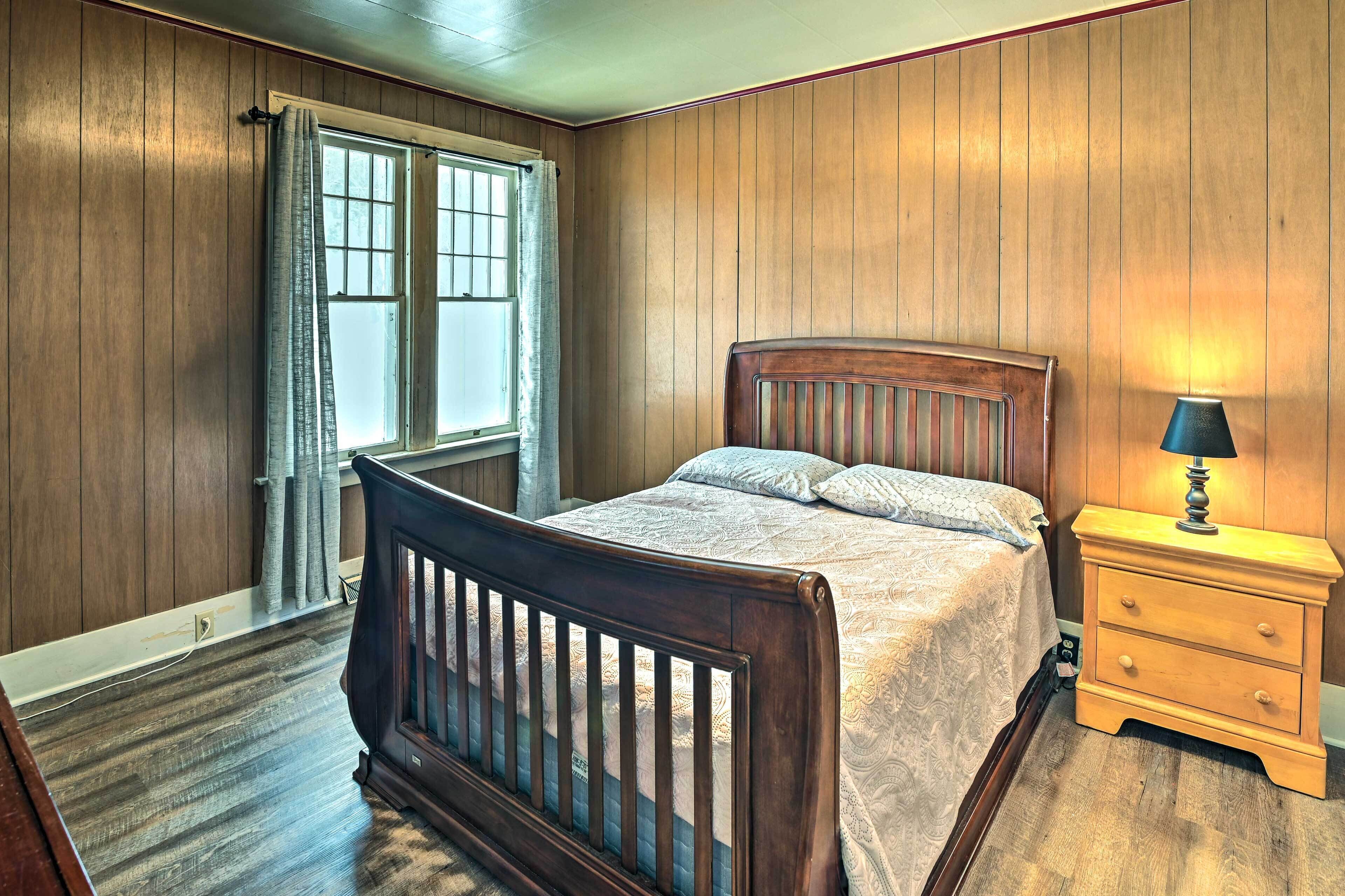 A bedroom with wooden paneled walls, a wooden bed frame, a nightstand with a lamp, and a window with curtains.