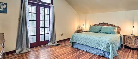 Une chambre avec un plancher en bois, un lit recouvert d’une courtepointe bleue, une table de chevet en bois, un ventilateur fixé au mur et une fenêtre avec des rideaux.