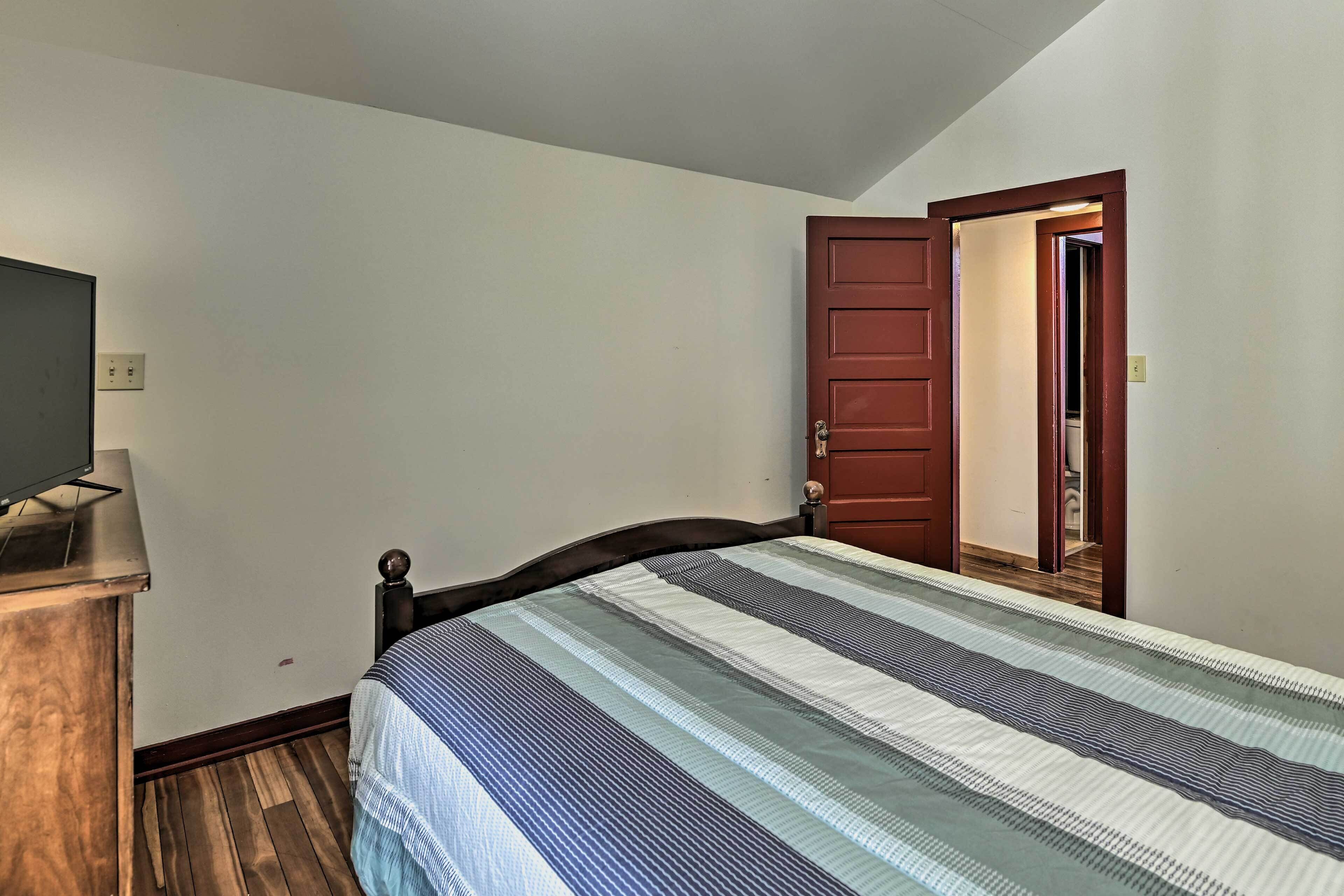 A bedroom with a bed, a wooden dresser, a television, and a door leading to another room.