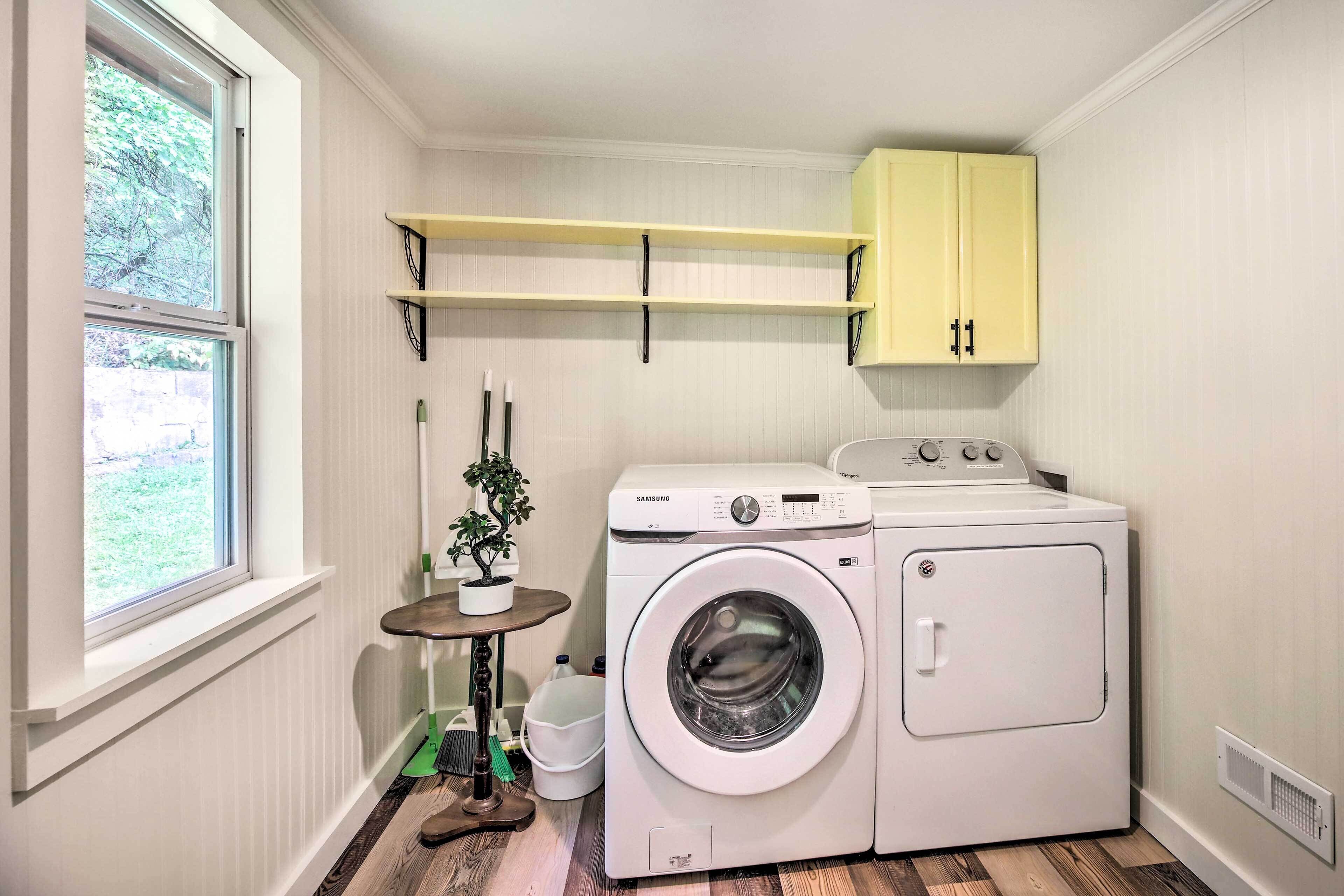 A laundry room with a washing machine, dryer, and a small table with a plant. There are shelves above the washing machine and a window on the left.
