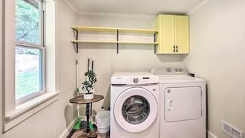 A laundry room with a washing machine, dryer, and a small table with a plant. There are shelves above the washing machine and a window on the left.
