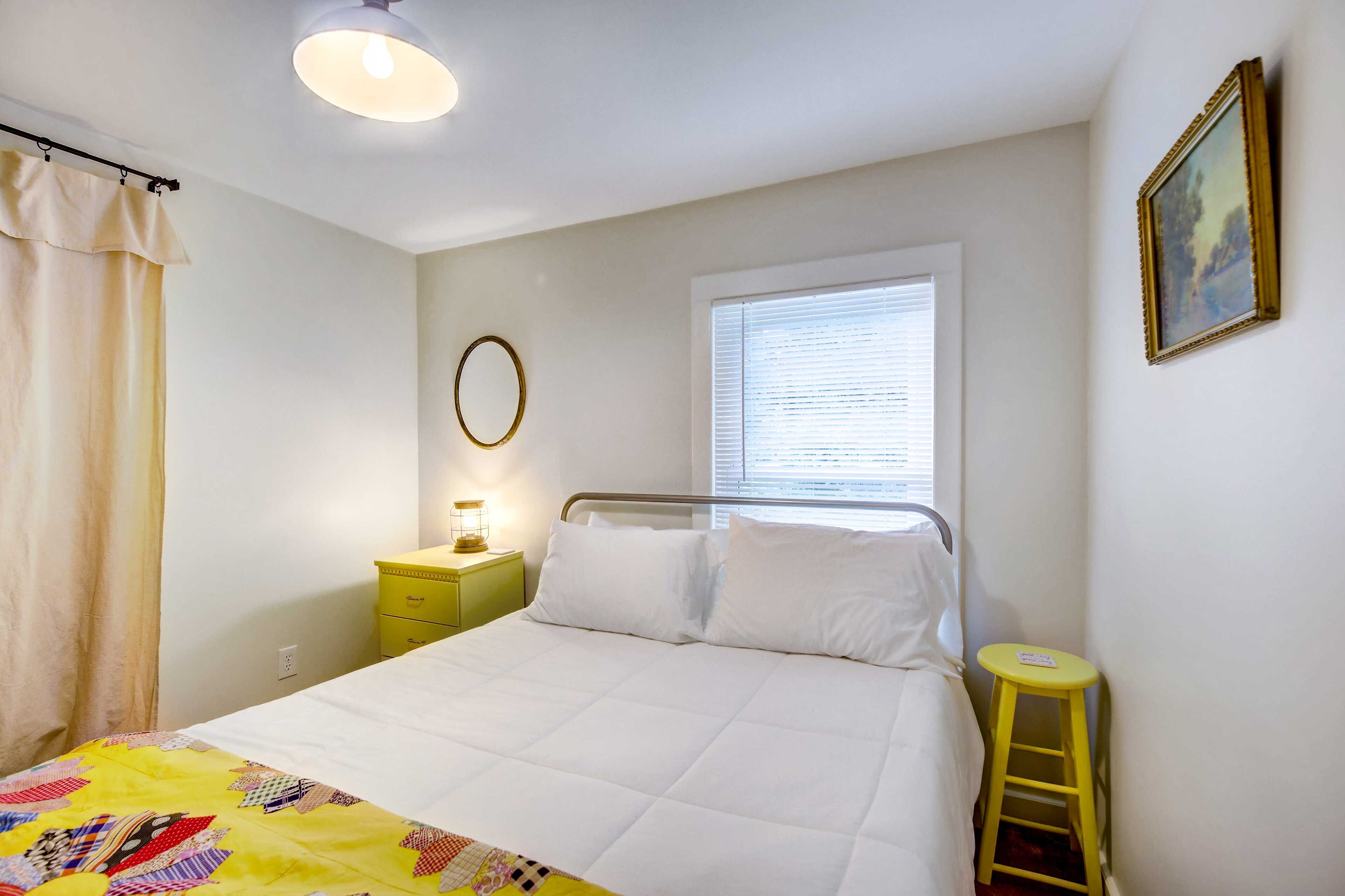 A bedroom with a bed, a nightstand, a window with blinds, a curtain, a wall-mounted picture, and a stool.