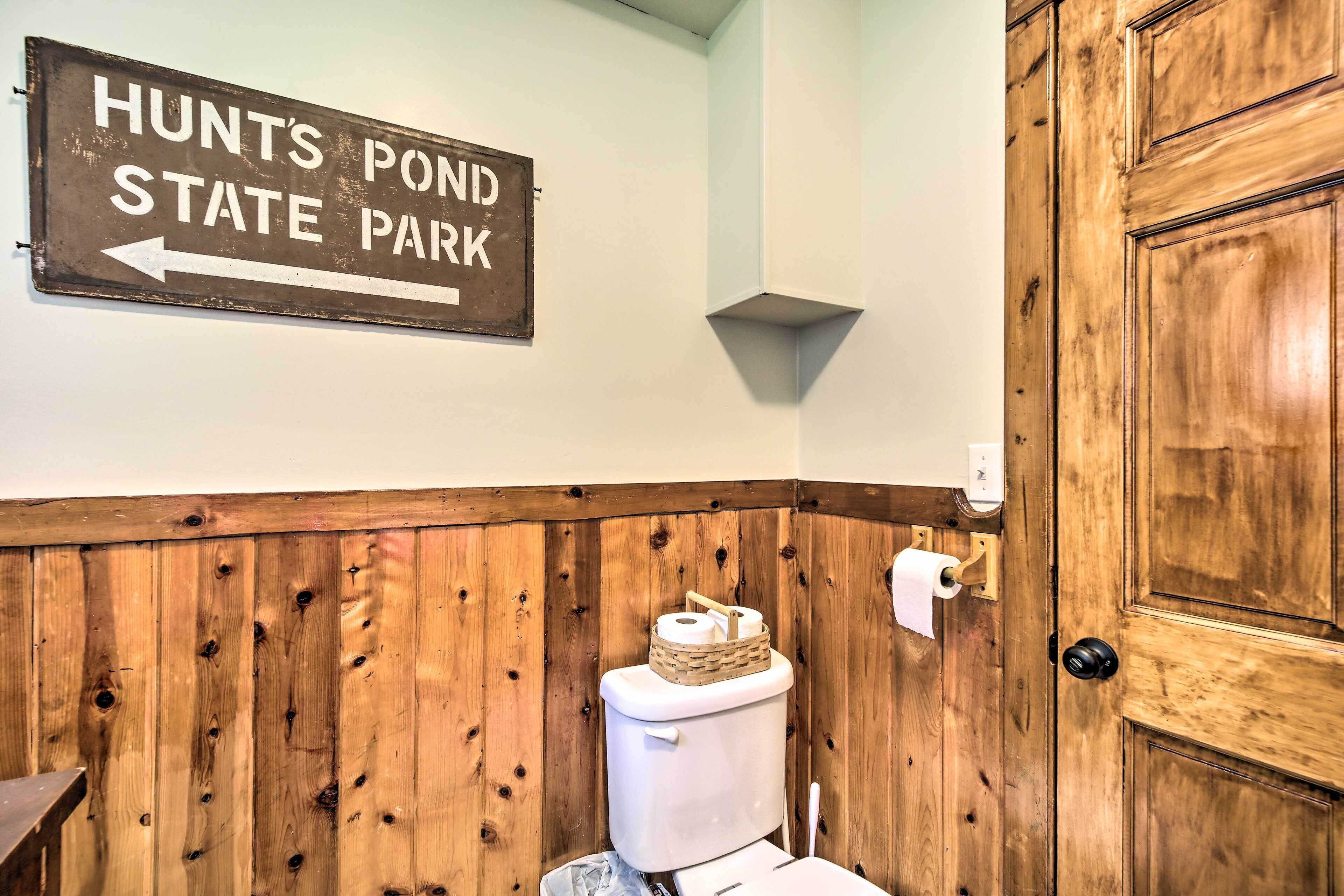 A bathroom with wooden paneling, a toilet, a sign indicating direction to Hunts Pond State Park, and a wooden door.