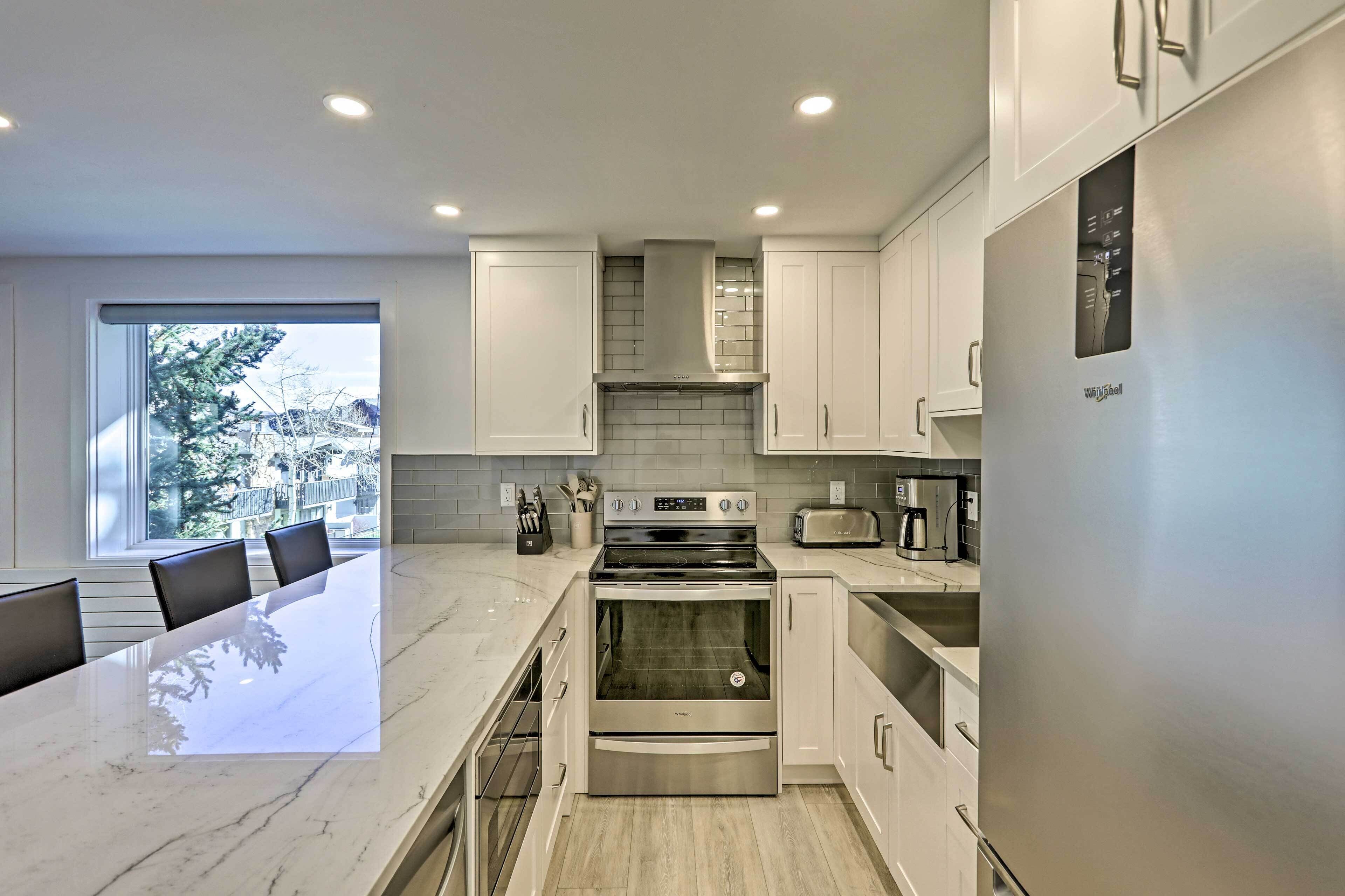 A modern kitchen with white cabinetry, a marble countertop, stainless steel appliances, and a view of trees through a large window.