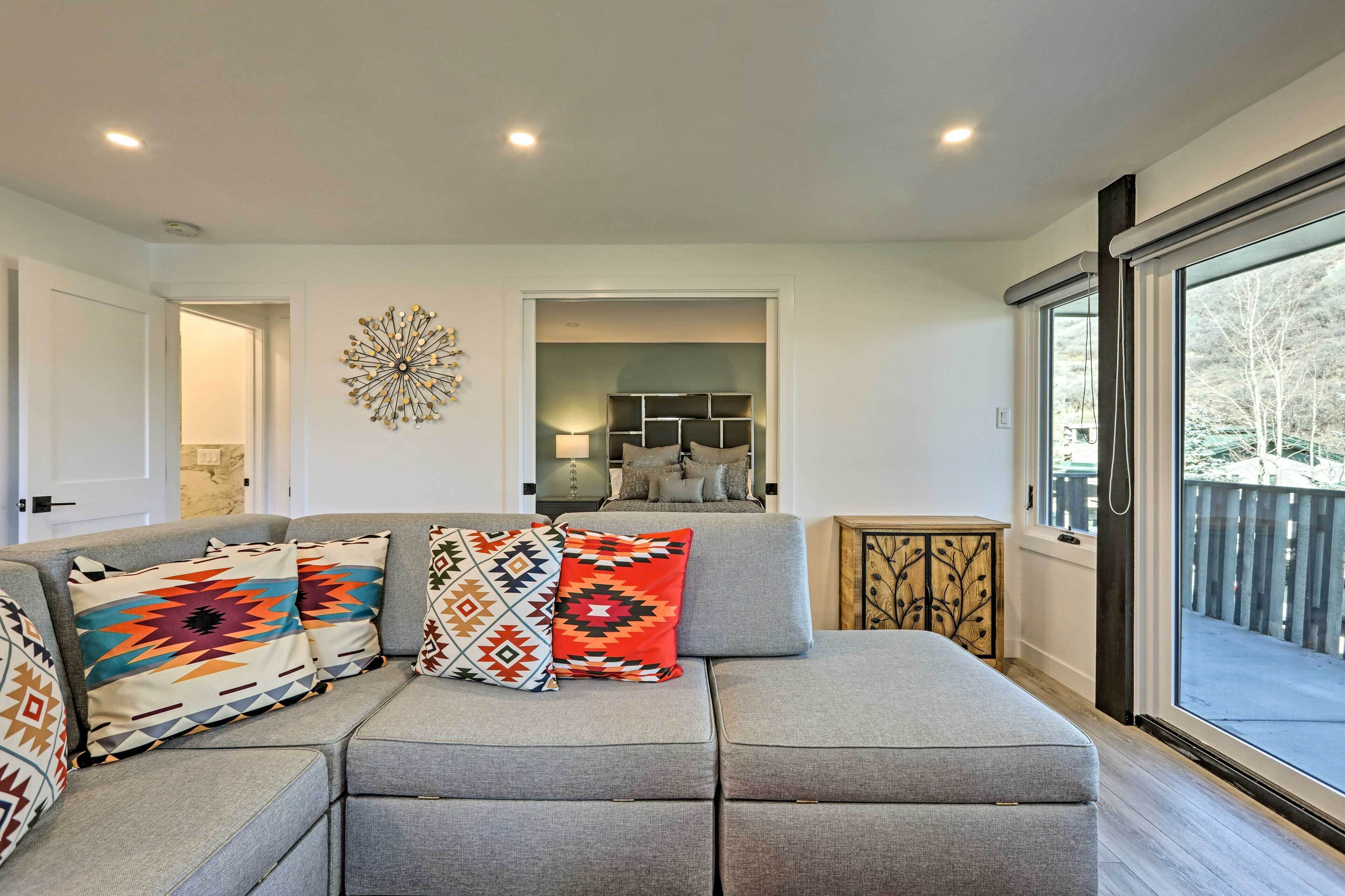 A modern living room with a grey sectional sofa, colorful throw pillows, a decorative wall piece, and a view through a large sliding glass door.