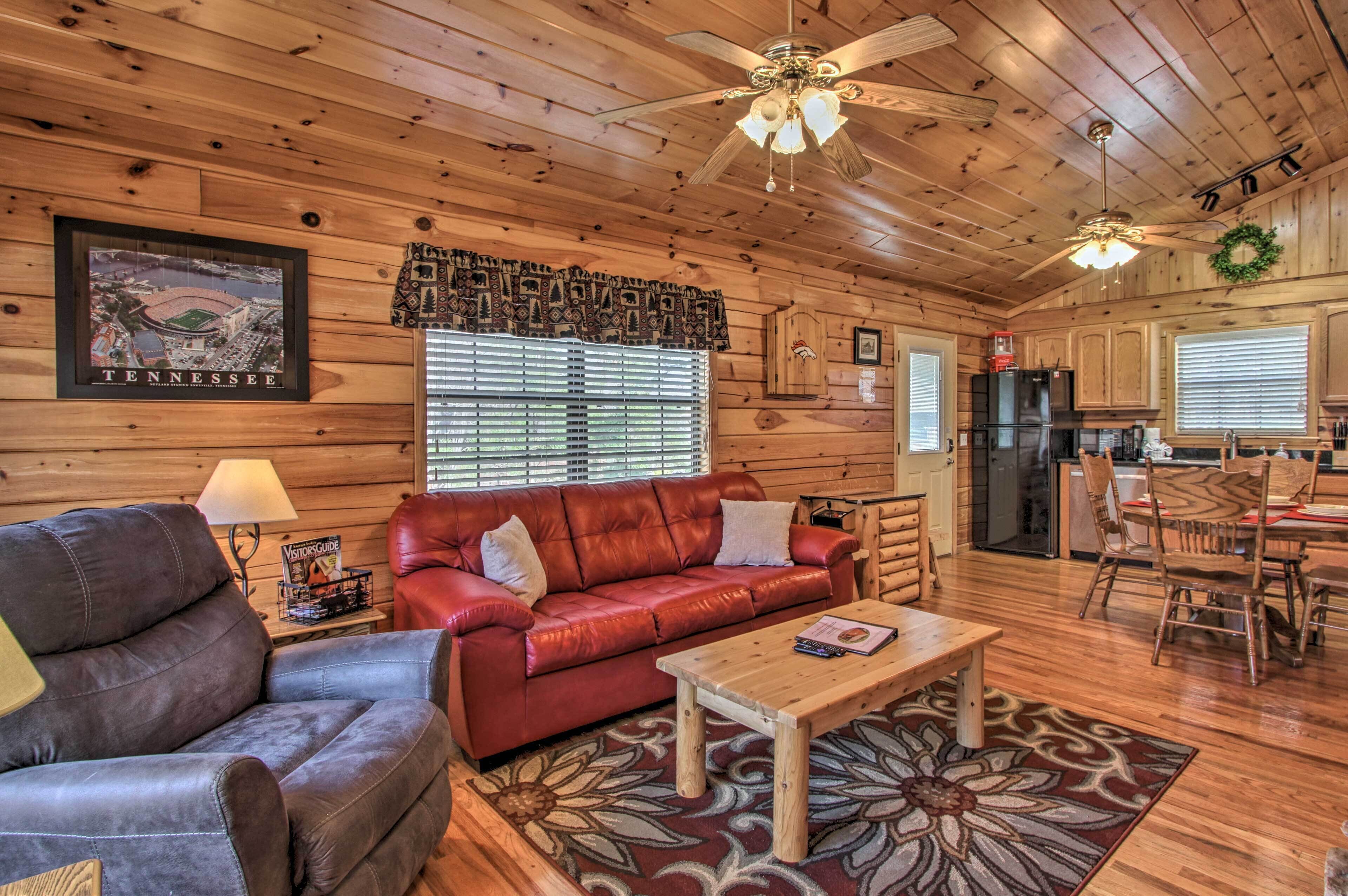 A cabin interior with a wooden ceiling, a red leather sofa, a wooden coffee table, and a dining area with a table and chairs.