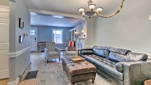 A living room with a sofa, ottoman, and chandelier. There is a dining area in the background with a table and chairs. - Historic Townhome in Downtown Shepherdstown! (Shepherdstown)