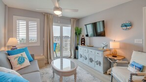 A living room with a ceiling fan, a flat-screen TV, a round clock, a glass coffee table, and a view of the outdoors through a sliding glass door. - Beach Access, Walk to Golf! Gulf Shores Condo (Gulf Shores)