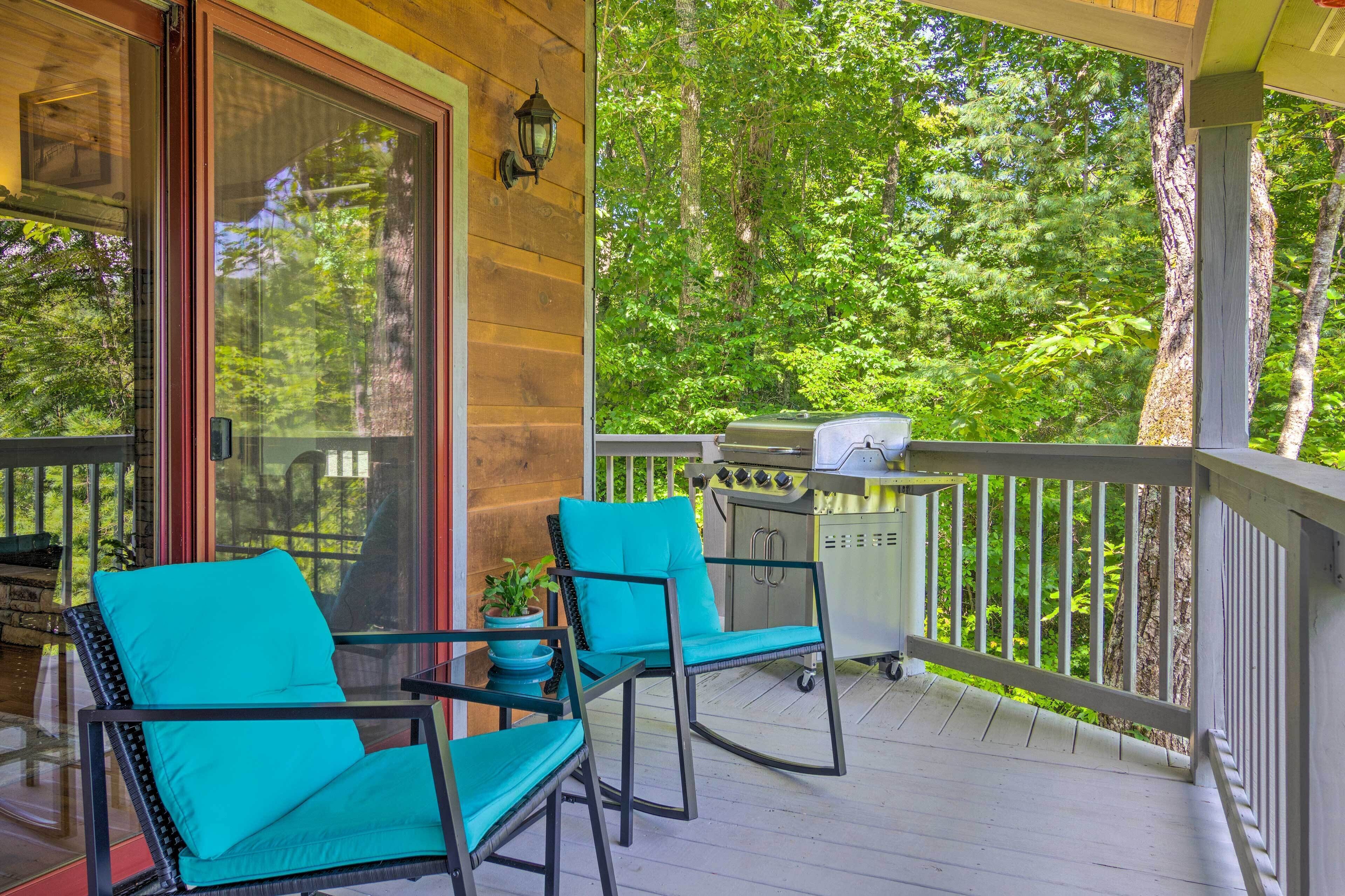 Eine überdachte Veranda mit zwei türkisfarbenen Stühlen, einem Grill und Blick auf ein bewaldetes Gebiet.