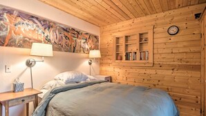 A wooden-paneled room with a bed, two bedside lamps, a wall clock, and a shelf with books. - Idyllic Northport Cottage, Walk to Lakeside! (Northport)