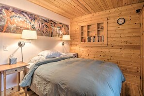 A wooden-paneled room with a bed, two bedside lamps, a wall clock, and a shelf with books. - Idyllic Northport Cottage, Walk to Lakeside! (Northport)