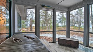 A covered porch with a bench and a small table, large windows offering a view of trees and a body of water. - Idyllic Northport Cottage, Walk to Lakeside! (Northport)