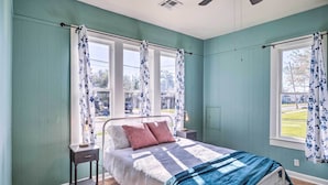 A bedroom with teal walls, a bed with a blue and white quilt, two bedside tables with lamps, and a window with floral curtains. - Lake Charles Cottage w/ Fireplace & Yard! (Lake Charles)