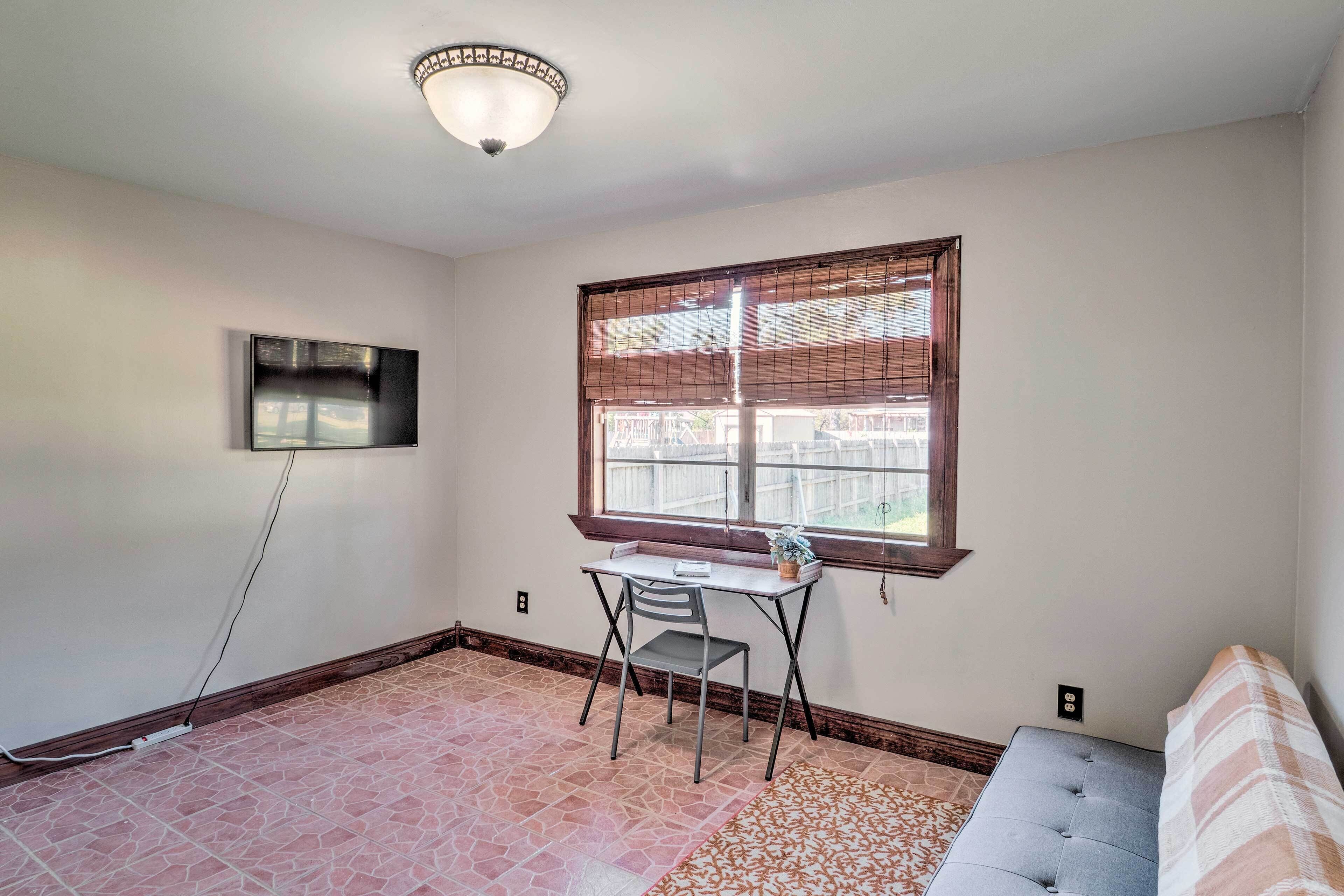 A room with a tiled floor, a desk with a chair, a sofa, a TV mounted on the wall, and a window with blinds.