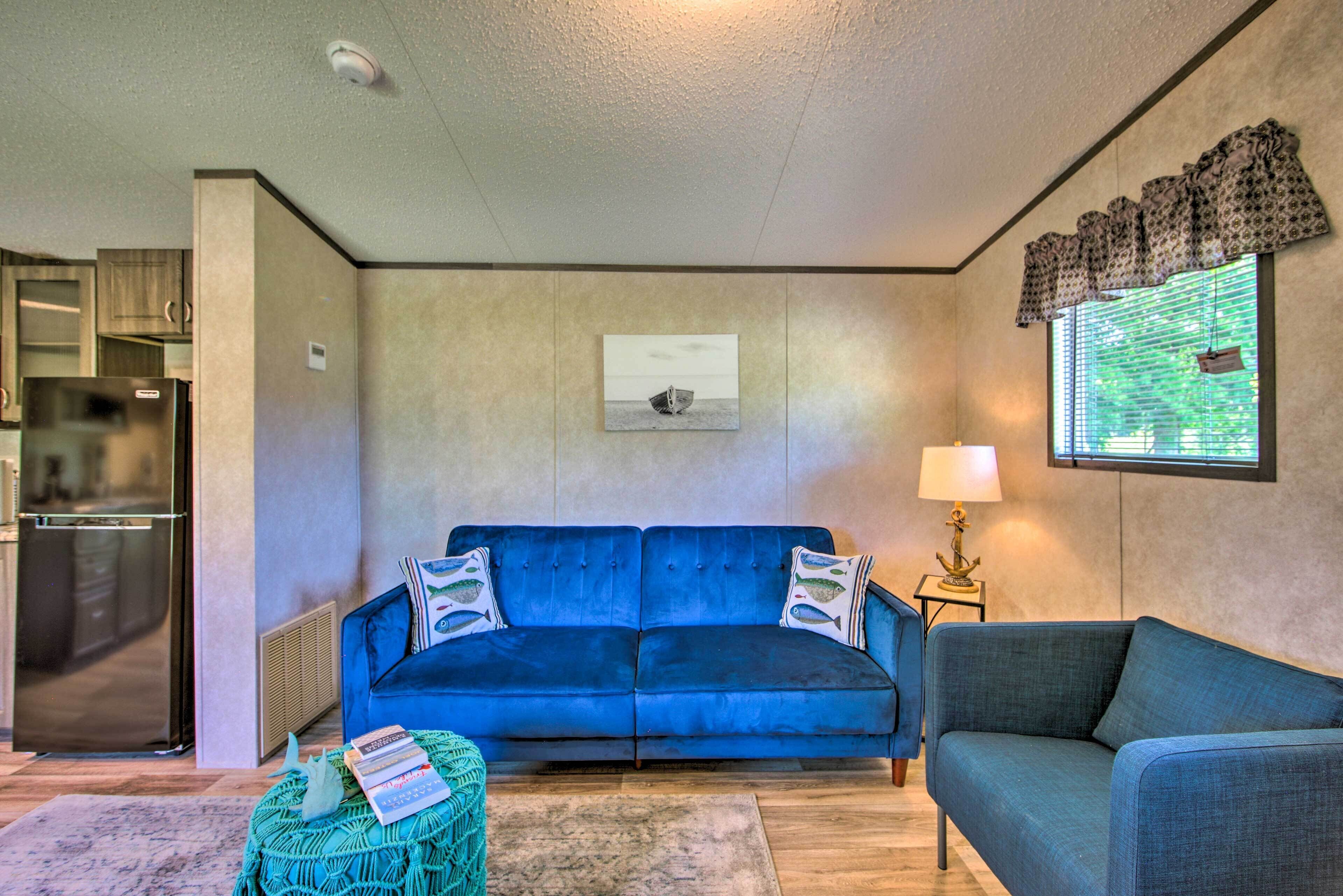 A living room with a blue sofa, a small table with a lamp, a framed picture on the wall, and a kitchen area with a refrigerator.