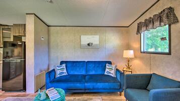 A living room with a blue sofa, a small table with a lamp, a framed picture on the wall, and a kitchen area with a refrigerator.