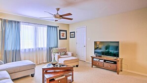 A living room with a ceiling fan, a TV on a wooden stand, a beige sofa, a coffee table, and a large window with curtains. - Home w/ Patio, Mins to Seaworld+lackland Afb! (San Antonio)