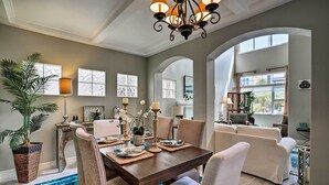A dining area with a wooden table, white chairs, and a chandelier. There are potted plants and a large window providing natural light. - Oceanside Home w/ Rooftop Patio, Walk to Beach! (Oceanside)