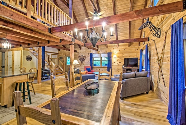 Interior de uma cabana de madeira com uma mesa de jantar, cadeiras, um sofá, uma televisão e uma escada.