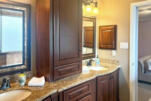 A bathroom with a granite countertop, two sinks, wooden cabinets, a mirror, and a towel. - Luxe Home 30 Miles to Houston w/ Pool & Grill (Cypress)