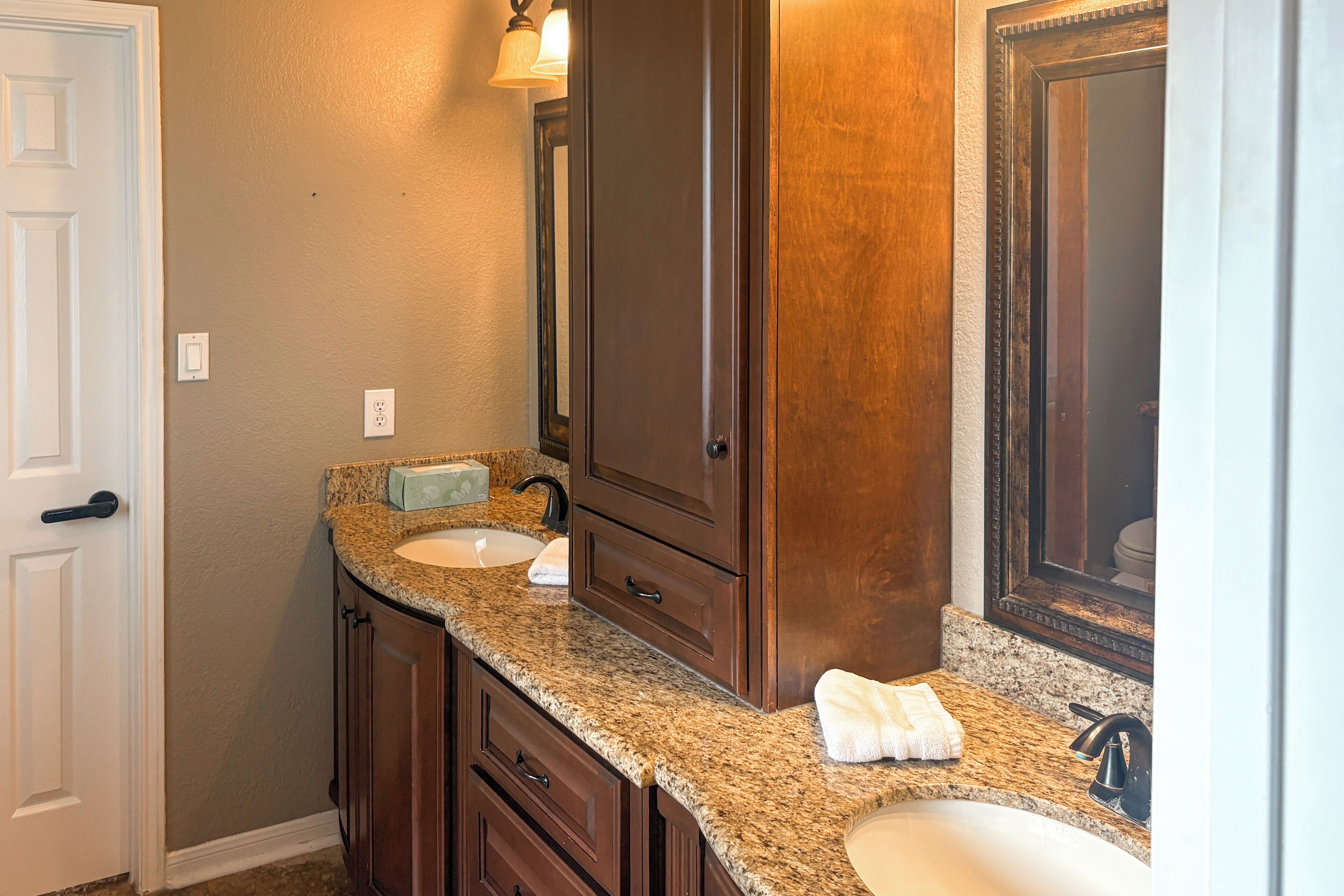 A bathroom with a granite countertop, two sinks, a mirror, and wooden cabinetry.