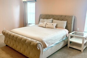 A bedroom with a tufted headboard, a large bed with a quilt, two bedside tables, and a window with blinds. - Luxe Home 30 Miles to Houston w/ Pool & Grill (Cypress)