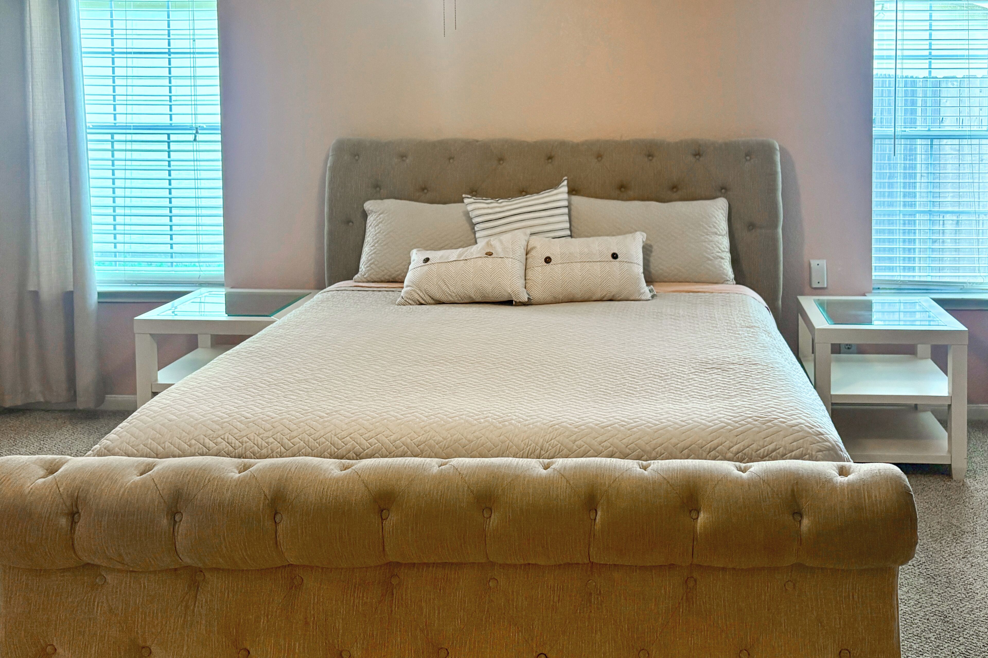 A bedroom with a tufted headboard, a large bed with a quilted bedspread, two bedside tables, and a window with blinds.