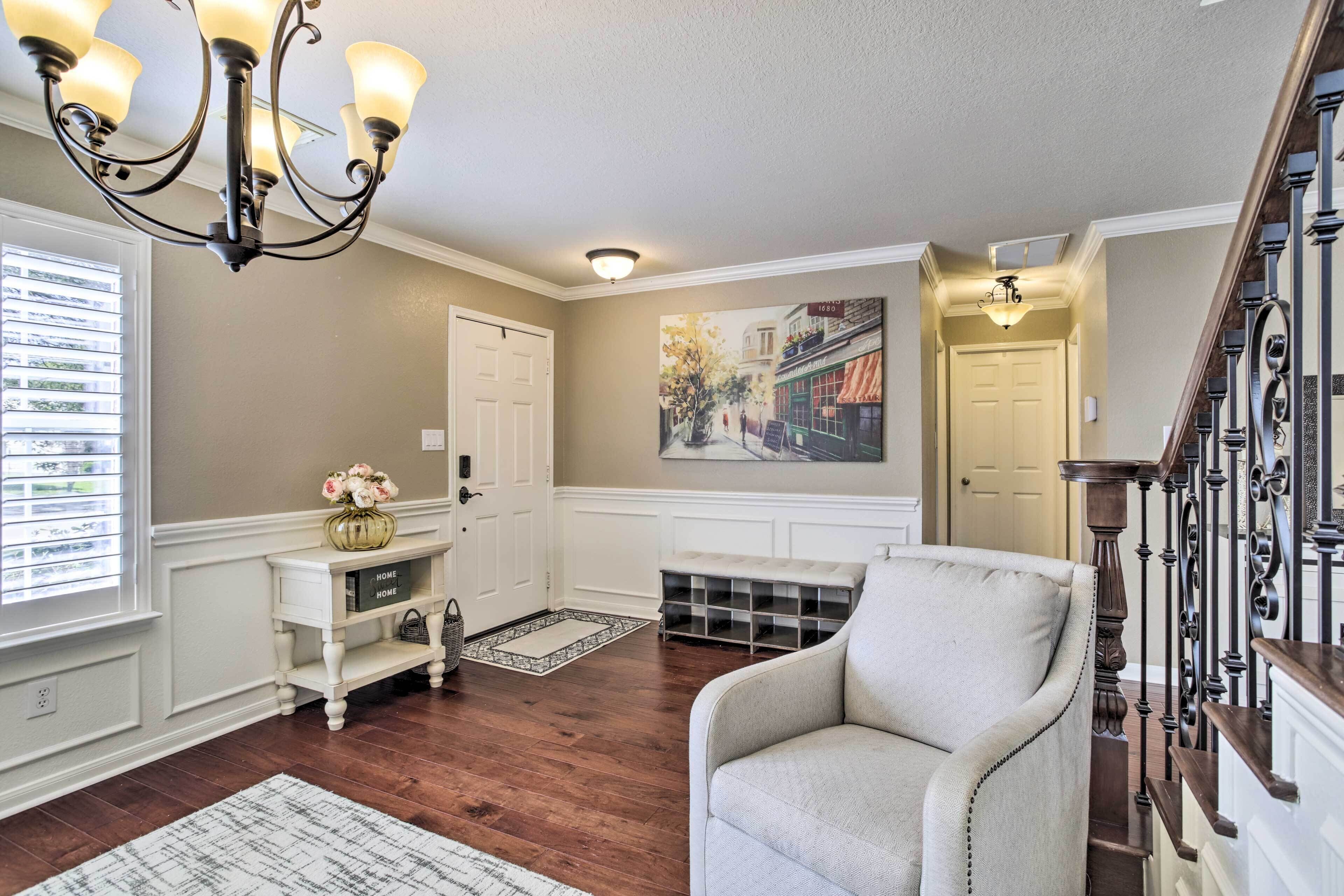 A living room with a chandelier, a white bench, a chair, a painting, and a staircase.