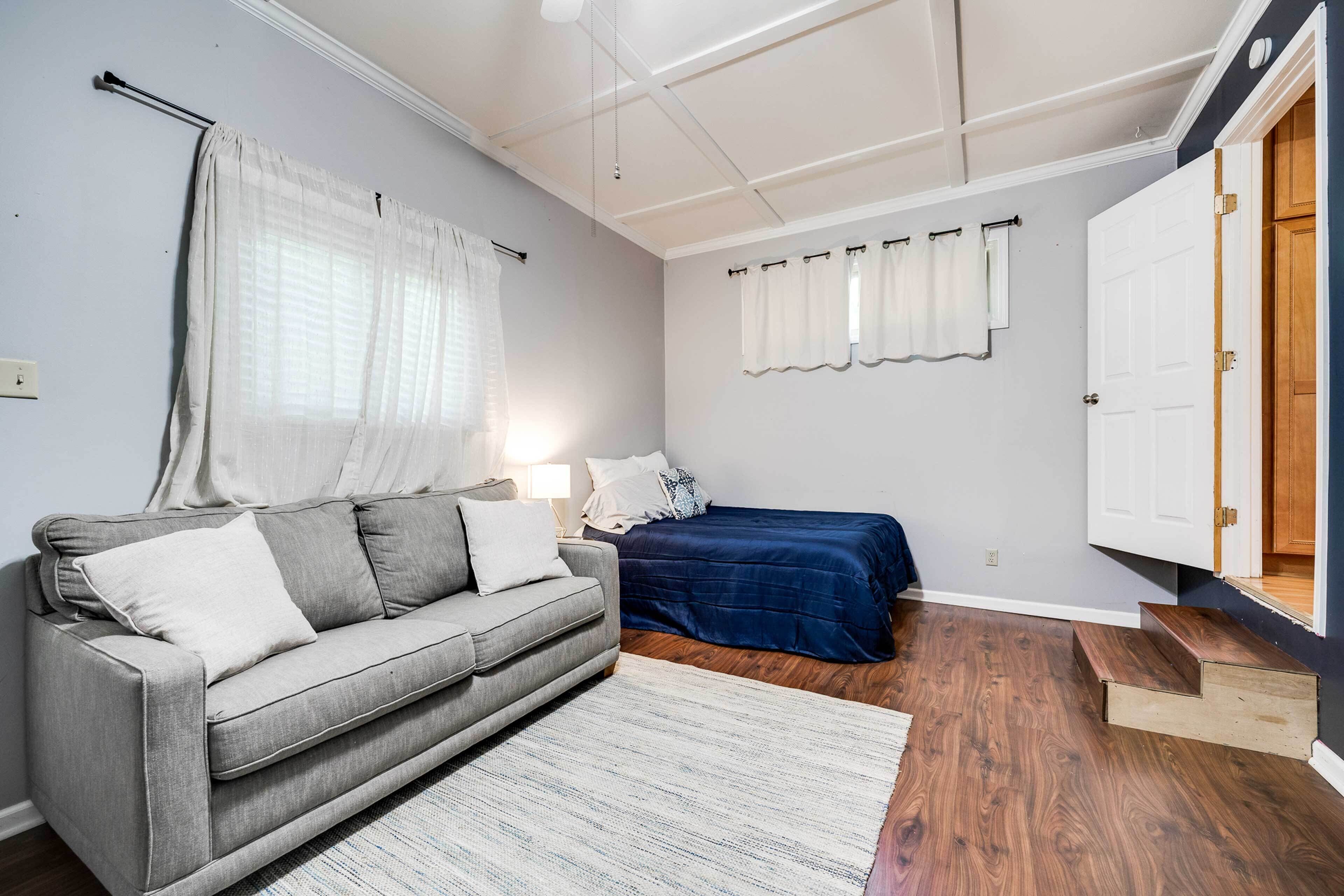 Um quarto com um sofá cinzento, uma cama com colcha azul, escada de madeira e uma porta branca.
