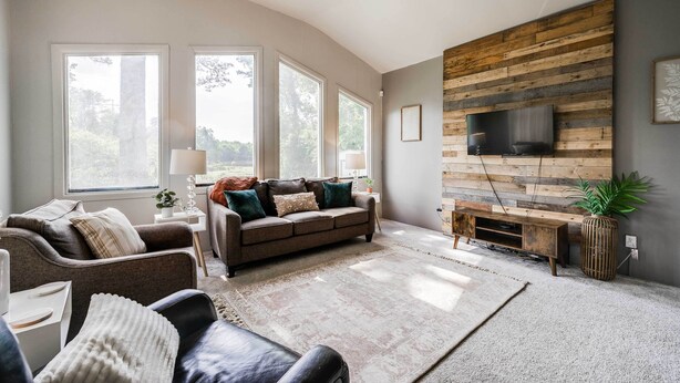Un salon moderne doté d’une grande fenêtre, d’un ventilateur de plafond, d’un mur d’accent en bois, d’un téléviseur et d’un canapé avec des coussins.