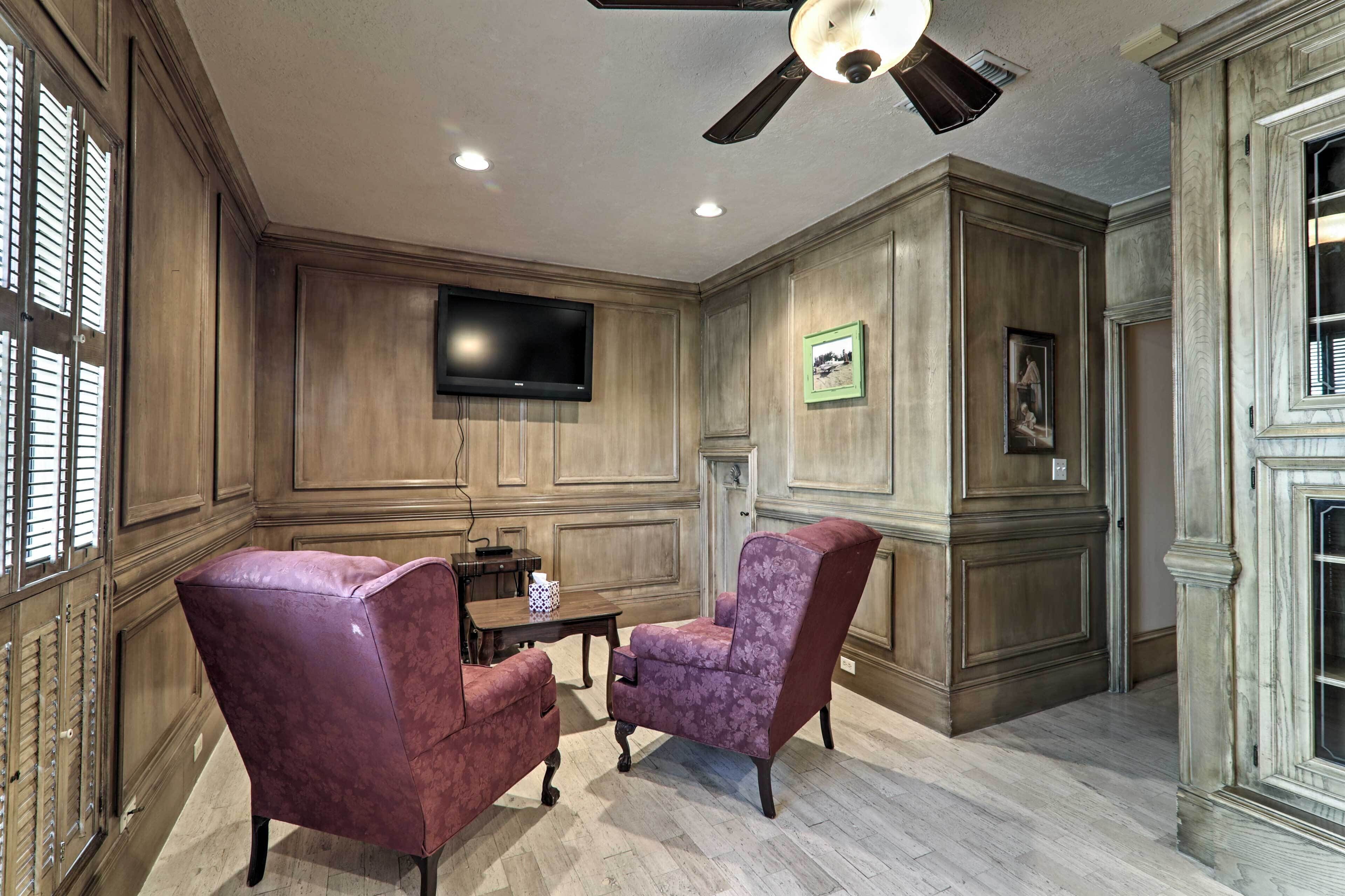 A room with wooden paneled walls, a ceiling fan, a flat-screen TV mounted on the wall, two upholstered armchairs, a small table with a decorative item, and a framed picture on the wall.