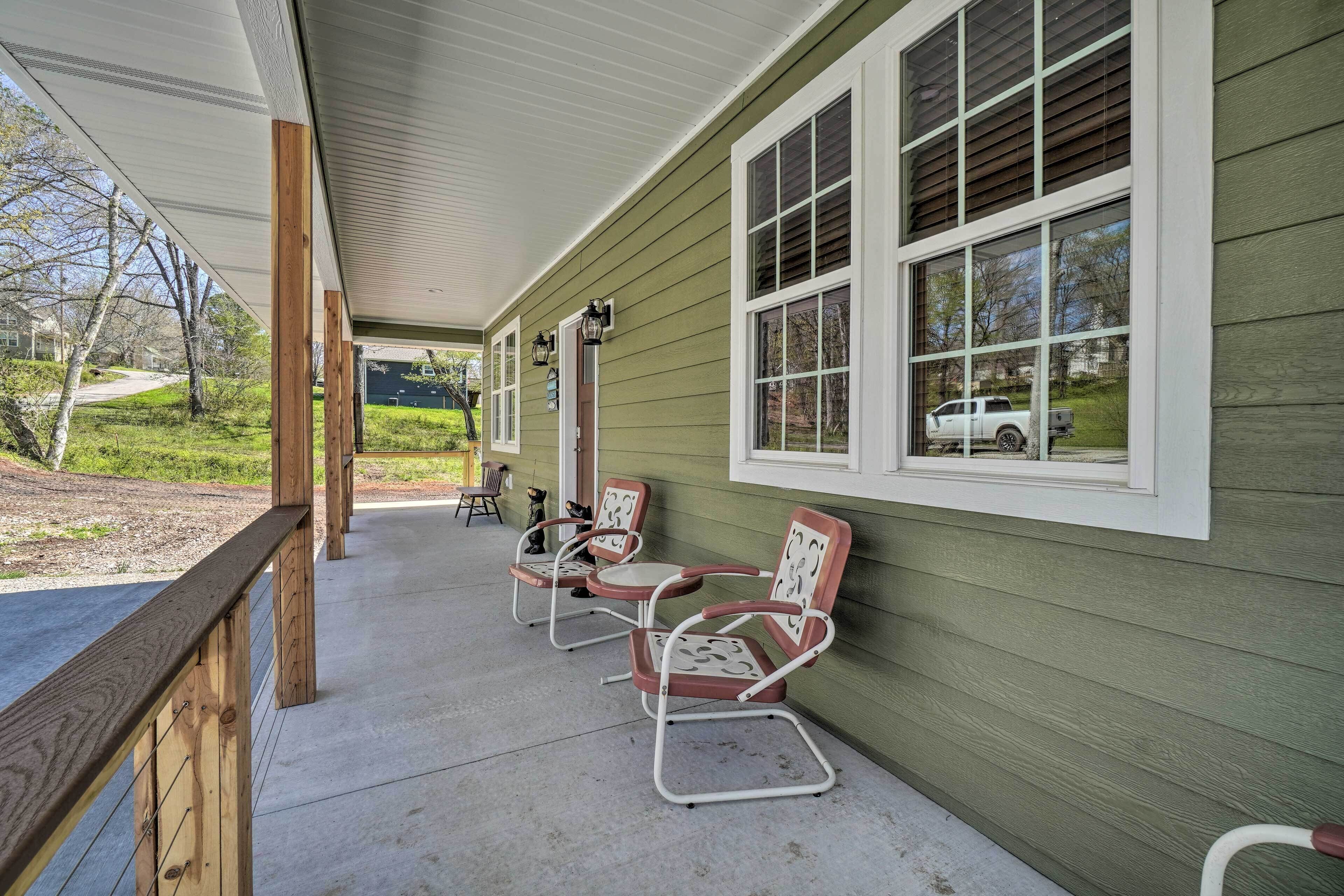 A covered porch with two chairs and a table, a wooden railing, and a view of a green house with large windows.