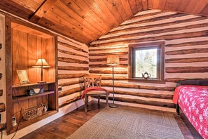 A log cabin room with a wooden floor, a bed with a red blanket, a chair, a lamp, a window with a view of trees, and a shelf with a lamp and a basket. - Macungie Cabin w/ Fireplace Near Bear Creek Skiing (Macungie)