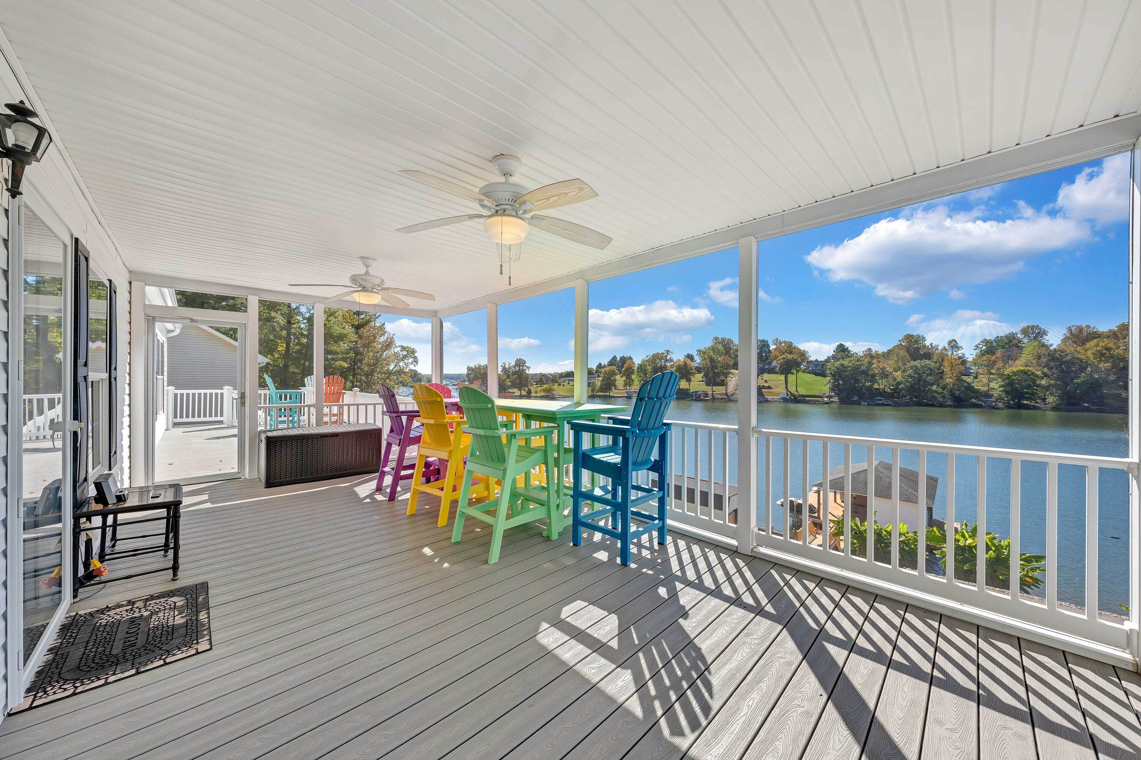 A covered deck with a view of a lake, featuring a table with colorful chairs and a ceiling fan.