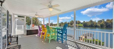 A covered deck with a view of a lake, featuring a table with colorful chairs and a ceiling fan.